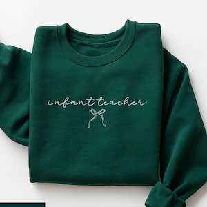 May include: Forest green sweatshirt featuring the text "infant teacher" in a cursive script, with a bow illustration. The garment is folded, displaying the crew neck and rolled-up sleeves. The colour is a deep, rich green.