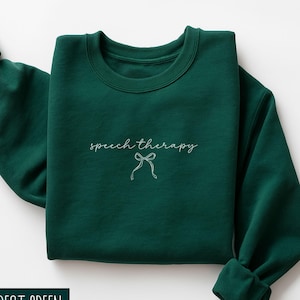 Peut inclure: Sweat-shirt vert forêt plié, avec l'inscription "speech therapy" en écriture cursive et un motif de nœud. Le texte blanc contraste avec le vert foncé. Le vêtement présente une texture douce et un design épuré.