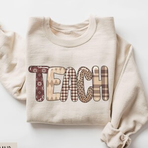 May include: A sand-coloured sweatshirt displaying the word "TEACH". The letters are designed with patchwork patterns, including plaid, leopard print, and geometric shapes in brown and beige. The sweatshirt has a crew neck and long sleeves.