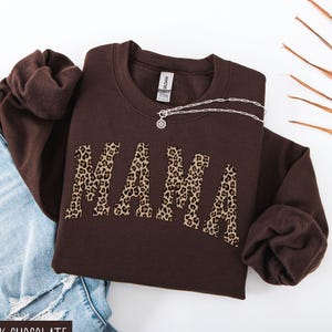 May include: A dark chocolate-colored sweatshirt with the word "MAMA" in leopard print across the front. A silver necklace rests on the sweatshirt. The sweatshirt is folded on top of a pair of blue jeans.