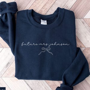 May include: A navy blue sweatshirt featuring the text "future mrs. johnson" in cursive, with a bow illustration. The sweatshirt has a round neck and long sleeves with ribbed cuffs. The word "NAVY" is printed in a white box in the bottom left corner.