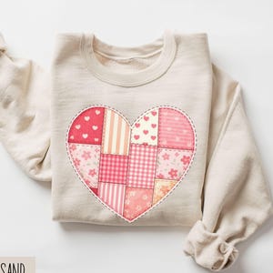May include: A sand-coloured sweatshirt with a large heart design. The heart is made up of pink and red patchwork squares with floral, striped, and heart patterns. The word "SAND" is printed at the bottom.