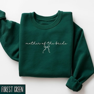 May include: A forest green sweatshirt featuring the phrase "mother of the bride" in cursive, with a bow illustration. The garment is neatly folded, highlighting its crew neck and long sleeves. The color is a deep, rich green.