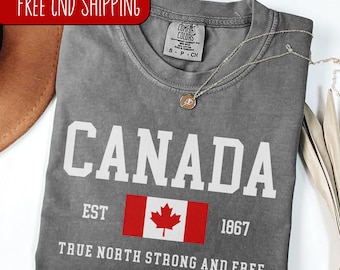 May include: A gray t-shirt with a white graphic of the word "CANADA" in a vintage style font. The graphic also includes the Canadian flag and the text "EST 1867 TRUE NORTH STRONG AND FREE".