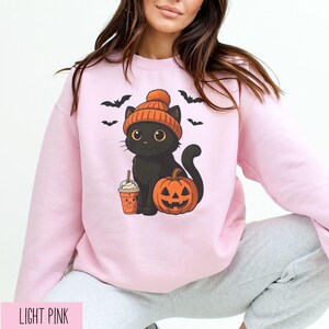 May include: Light pink sweatshirt featuring a black cat wearing an orange beanie, holding a pumpkin spice drink, and sitting next to a jack-o'-lantern. Black bats are flying above the cat. The text "LIGHT PINK" is at the bottom of the image.