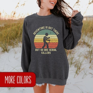 May include: A dark grey sweatshirt with a vintage-style graphic that says "Adventure is out there, but so are serial killers". The graphic features a stick figure hiking with a hiking stick in front of a sunset.