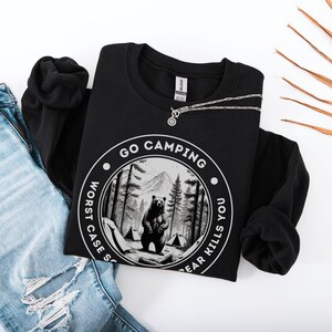 Puede incluir: Sudadera negra con un gr&aacute;fico circular blanco que dice "Go Camping Worst Case Scenario Bear Kills You". El gr&aacute;fico presenta un oso negro de pie en un bosque con una monta&ntilde;a en el fondo.