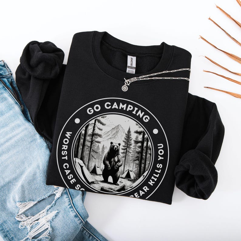 Puede incluir: Sudadera negra con un gr&aacute;fico circular blanco que dice "Go Camping Worst Case Scenario Bear Kills You". El gr&aacute;fico presenta un oso negro de pie en un bosque con una monta&ntilde;a en el fondo.