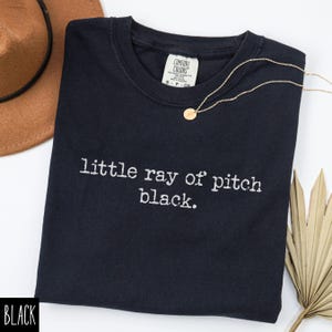 May include: Black crew neck t-shirt with the phrase "little ray of pitch black" in white lettering. A gold necklace with a small pendant rests on the shirt. A brown hat and dried palm leaf are in the background.