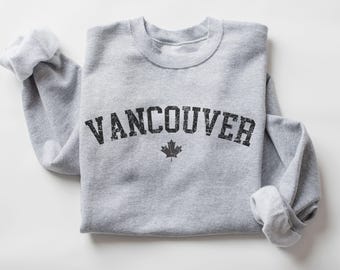 May include: A heather gray sweatshirt with the word "VANCOUVER" in distressed black lettering and a black maple leaf graphic. The sleeves are rolled up, and the sweatshirt is folded neatly.