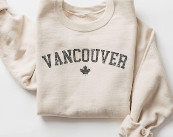 May include: A cream-colored sweatshirt with the word "VANCOUVER" printed in distressed gray lettering. A small gray maple leaf is centered below the text. The sleeves are folded up, and the fabric appears soft and comfortable.