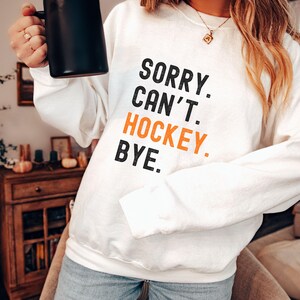 May include: White sweatshirt with the text "SORRY. CAN'T. HOCKEY. BYE." in black and orange lettering.