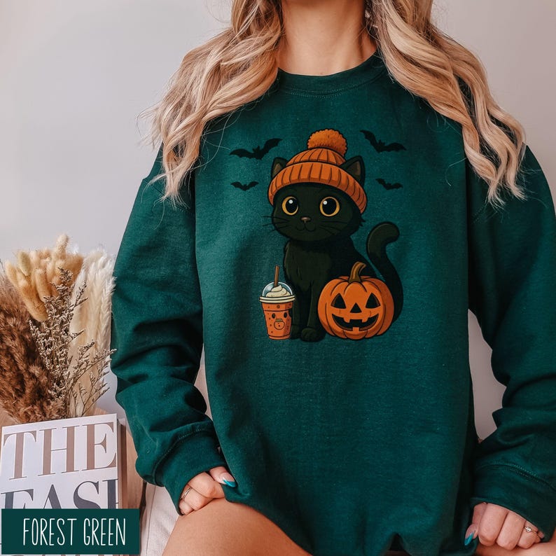 May include: Forest green sweatshirt featuring a black cat wearing an orange beanie, holding a drink, and sitting next to a carved pumpkin. The design includes bats and is perfect for Halloween. The text "FOREST GREEN" is at the bottom.