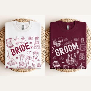 May include: Two white t-shirts with red ink designs. The left shirt says "Bride" and has wedding-themed illustrations. The right shirt says "Groom" and has groom-themed illustrations.