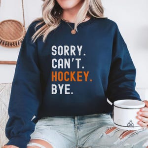 May include: A navy blue sweatshirt with the text "SORRY. CAN'T. HOCKEY. BYE." in white and orange lettering.
