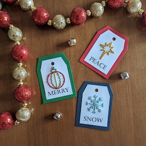 Peut inclure: Trois étiquettes cadeaux de Noël avec des motifs brodés. Les étiquettes sont rouge, verte et bleue avec du texte blanc. Les motifs sont une étoile, un ornement et un flocon de neige. Le texte sur les étiquettes indique "Paix", "Joyeux Noël" et "Neige".