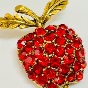 Puede incluir: Broche vintage de fresa dorada, con tallo y hojas. La fresa está incrustada con numerosos pedrería roja. El broche tiene un diseño clásico y elegante, adecuado para añadir un toque de brillo a cualquier atuendo.