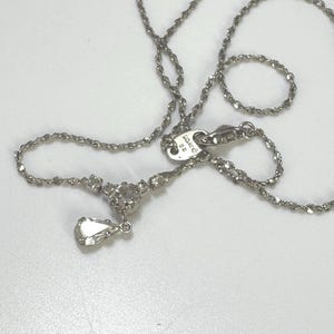 Könnte beinhalten: Eine silberne Halskette mit zarter Kette und einem Anhänger mit einem tropfenförmigen Edelstein. Die Halskette hat eine kleine Ansammlung von Edelsteinen in der Nähe des Anhängers und einen Verschluss. Der Schmuck wird auf einer weißen Oberfläche präsentiert.