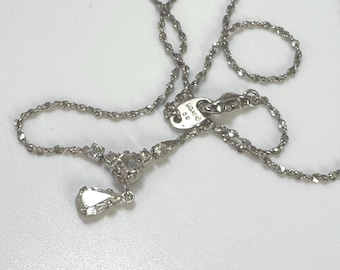 Op de afbeelding: Een zilveren ketting met een fijne ketting en een hanger met een druppelvormige edelsteen. De ketting heeft een kleine cluster van edelstenen bij de hanger en een sluiting. De sieraden worden gepresenteerd op een wit oppervlak.