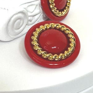 May include: A pair of red button earrings with a gold rope-like border. The earrings have a glossy finish and a round shape. The earrings are set against a white background.