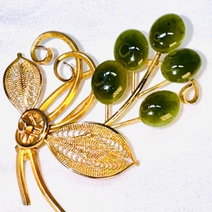May include: A gold-tone metal brooch featuring two filigree leaves and swirling wire accents paired with a cluster of five smooth, oval-shaped dark green stones. The design resembles a botanical spray with polished metal stems and textured foliage.