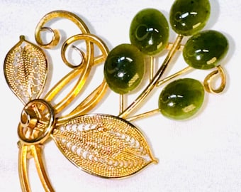 May include: A gold-tone metal brooch featuring two filigree leaves and swirling wire accents paired with a cluster of five smooth, oval-shaped dark green stones. The design resembles a botanical spray with polished metal stems and textured foliage.