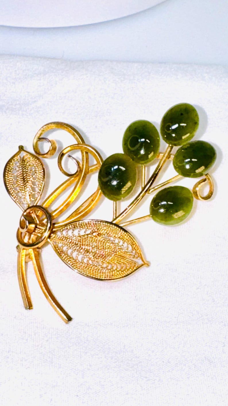May include: A gold-tone metal brooch featuring two filigree leaves and swirling wire accents paired with a cluster of five smooth, oval-shaped dark green stones. The design resembles a botanical spray with polished metal stems and textured foliage.