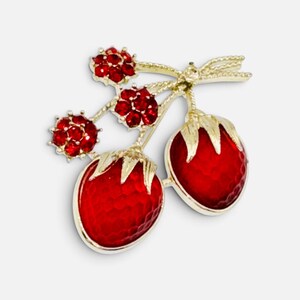May include: A gold-tone brooch shaped like two strawberries with textured red enamel bodies and gold leaves. The stems are adorned with three small, round, red rhinestone-encrusted flower accents, creating a detailed fruit-inspired accessory.