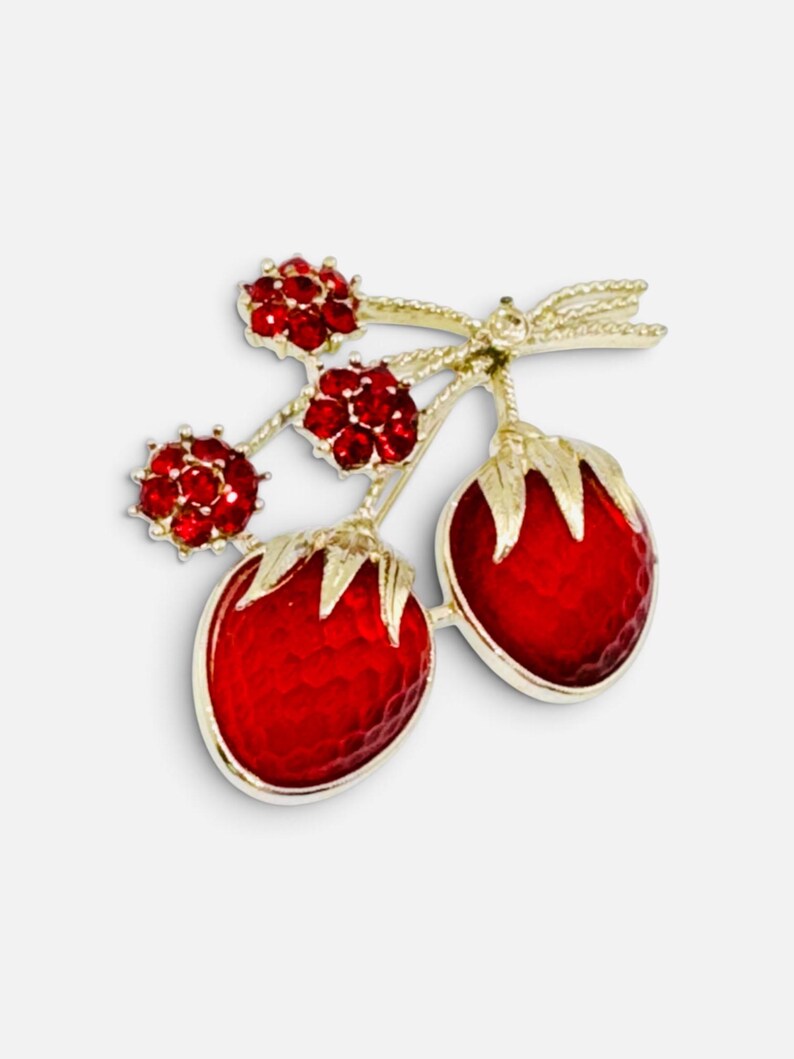 May include: A gold-tone brooch shaped like two strawberries with textured red enamel bodies and gold leaves. The stems are adorned with three small, round, red rhinestone-encrusted flower accents, creating a detailed fruit-inspired accessory.