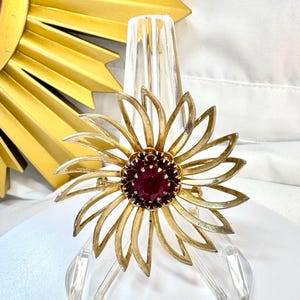 Op de afbeelding: Een goudkleurige metalen broche in de vorm van een bloem met langwerpige, puntige bloemblaadjes die uitstralen vanuit een centrum van dieprode, gefacetteerde ronde stenen. De broche heeft een textuur op de blaadjes en een speld.