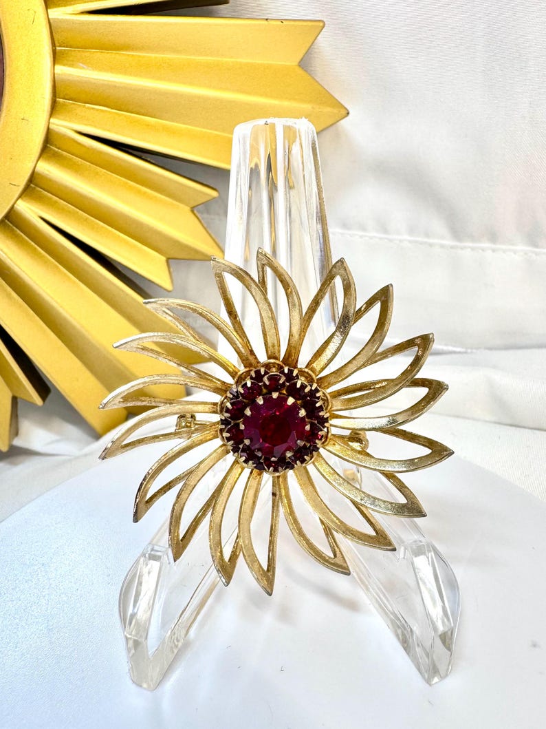 Op de afbeelding: Een goudkleurige metalen broche in de vorm van een bloem met langwerpige, puntige bloemblaadjes die uitstralen vanuit een centrum van dieprode, gefacetteerde ronde stenen. De broche heeft een textuur op de blaadjes en een speld.