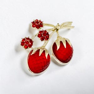 May include: A gold-tone brooch shaped like two strawberries with textured red enamel bodies and gold leaves. The stems are adorned with three small clusters of sparkling red rhinestones, creating a detailed fruit-inspired accessory.