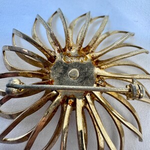 Op de afbeelding: Een goudkleurige metalen broche in de vorm van een bloem met dunne, uitstralende bloemblaadjes. Het midden is voorzien van een ronde plaat met de tekst "SARAH COV" gestempeld. Een horizontale speldbevestiging zit aan de achterkant.