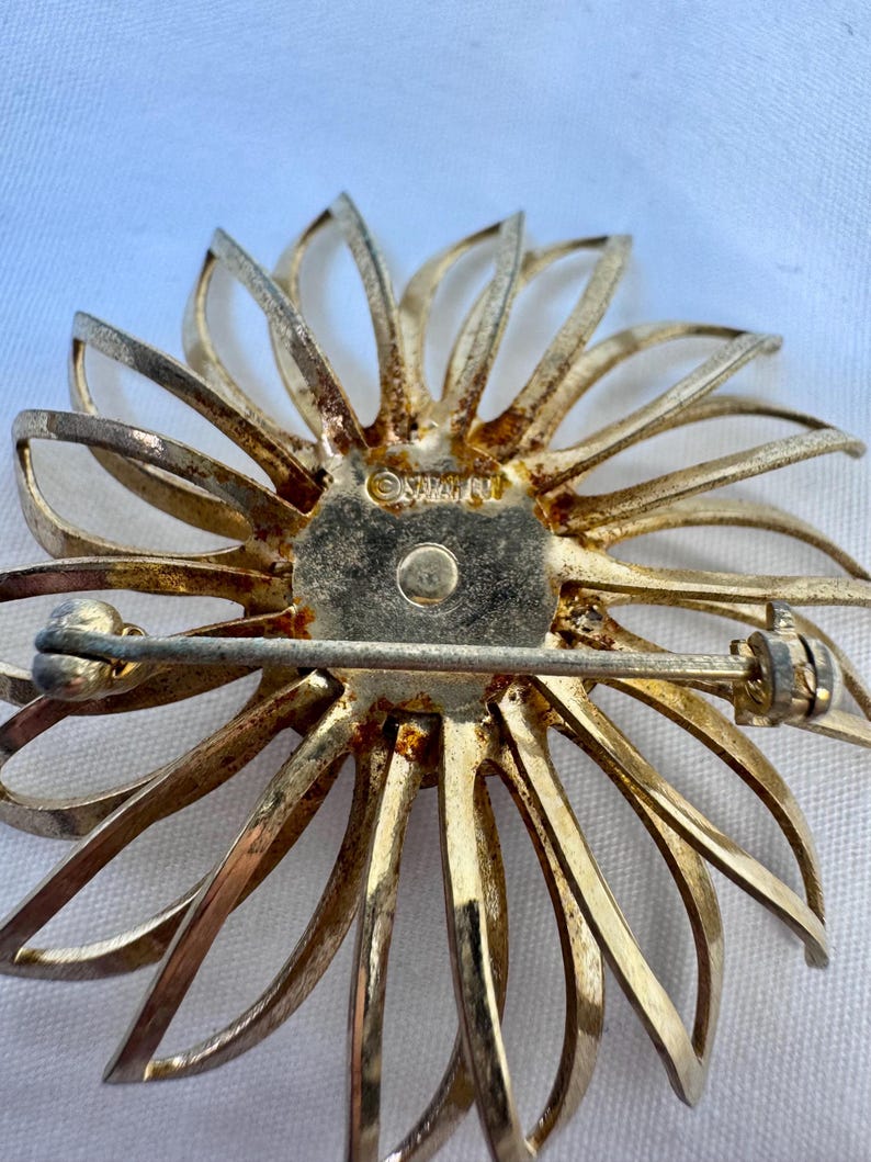Op de afbeelding: Een goudkleurige metalen broche in de vorm van een bloem met dunne, uitstralende bloemblaadjes. Het midden is voorzien van een ronde plaat met de tekst "SARAH COV" gestempeld. Een horizontale speldbevestiging zit aan de achterkant.