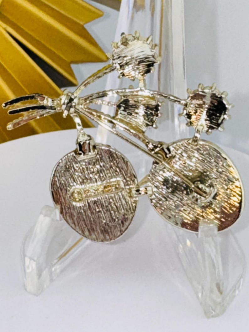 May include: A silver-toned metal brooch featuring a cluster of three circular elements attached to stems. The back of the brooch displays a textured finish with a horizontal pin mechanism and a clearly visible stamped hallmark on the left side.