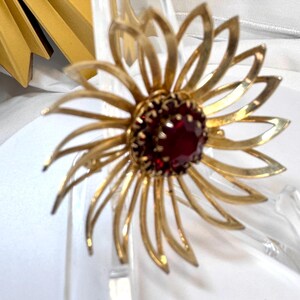 Op de afbeelding: Een goudkleurige metalen broche in de vorm van een bloem met langwerpige, puntige bloemblaadjes die uitstralen vanaf een centrale, ronde donkerrode edelsteen. De broche heeft een gepolijste afwerking en een klauwzetting voor de steen.