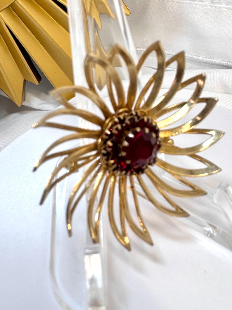 Op de afbeelding: Een goudkleurige metalen broche in de vorm van een bloem met langwerpige, puntige bloemblaadjes die uitstralen vanaf een centrale, ronde donkerrode edelsteen. De broche heeft een gepolijste afwerking en een klauwzetting voor de steen.