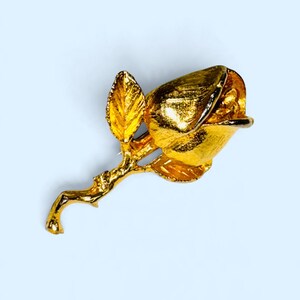 K&ouml;nnte beinhalten: Eine goldfarbene Metallbrosche in Form einer Rosenknospe an einem Stiel mit zwei Bl&auml;ttern. Die Oberfl&auml;che des Metalls weist eine texturierte, geb&uuml;rstete Optik auf, welche die Rundungen der Bl&uuml;tenbl&auml;tter und die organische Form des Stiels betont.