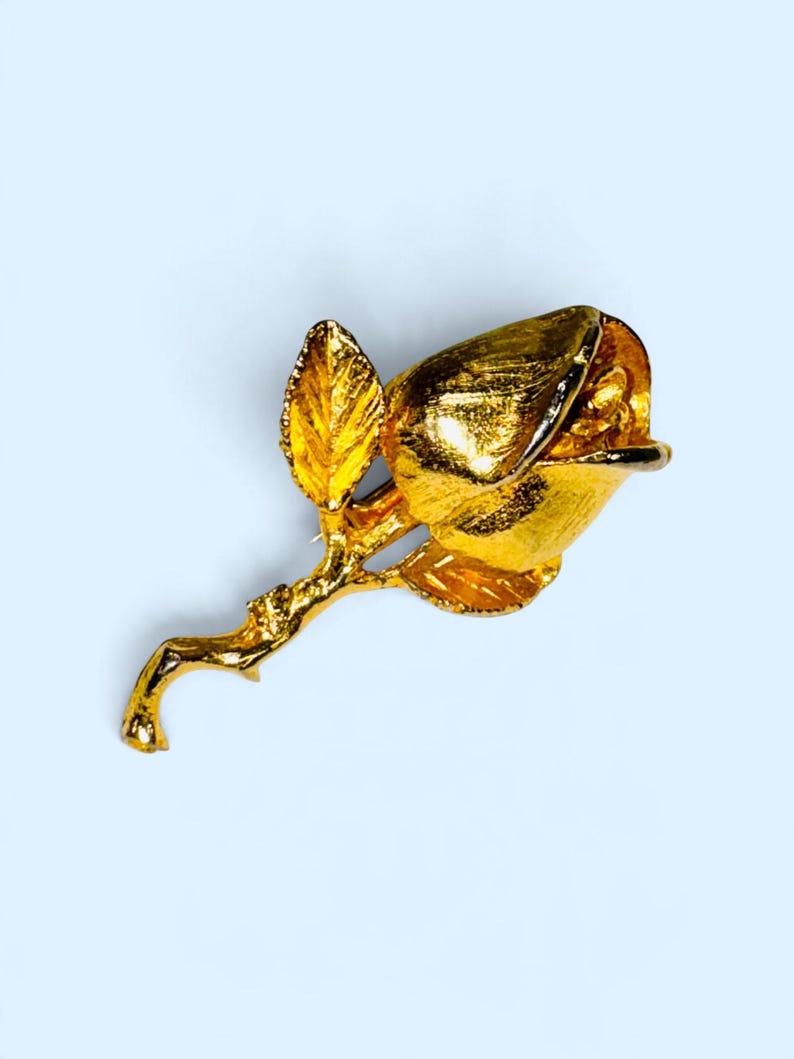 K&ouml;nnte beinhalten: Eine goldfarbene Metallbrosche in Form einer Rosenknospe an einem Stiel mit zwei Bl&auml;ttern. Die Oberfl&auml;che des Metalls weist eine texturierte, geb&uuml;rstete Optik auf, welche die Rundungen der Bl&uuml;tenbl&auml;tter und die organische Form des Stiels betont.