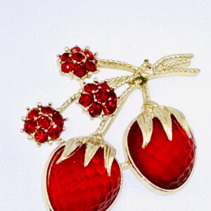 May include: A gold-tone brooch shaped like two strawberries with red textured enamel bodies and gold leaves, accented by three small flower-shaped clusters of red rhinestones on gold stems, all connected by a gold-tone bow detail.