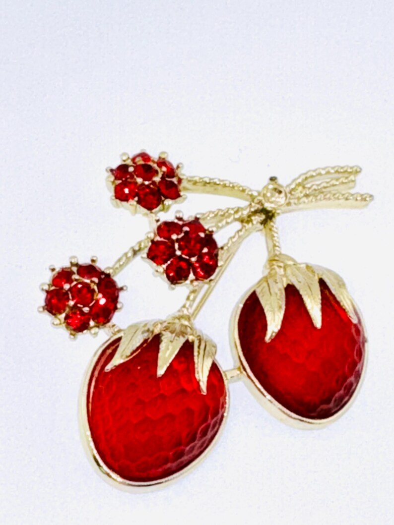 May include: A gold-tone brooch shaped like two strawberries with red textured enamel bodies and gold leaves, accented by three small flower-shaped clusters of red rhinestones on gold stems, all connected by a gold-tone bow detail.
