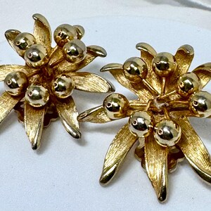May include: A pair of gold-toned metal earrings featuring a textured, leaf-like base with multiple smooth, spherical beads clustered in the center. The design mimics a floral arrangement with brushed metal petals radiating outward from the center.