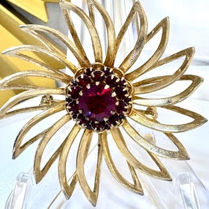 Op de afbeelding: Een goudkleurige metalen broche in de vorm van een bloem met langwerpige, opengewerkte bloemblaadjes. Het midden van de bloem bevat een grote, ronde, dieprode gefacetteerde steen omringd door een ring van kleinere, bijpassende rode stenen.