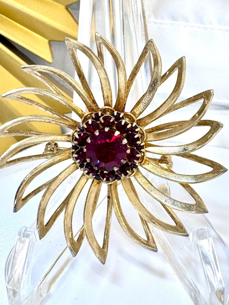 Op de afbeelding: Een goudkleurige metalen broche in de vorm van een bloem met langwerpige, opengewerkte bloemblaadjes. Het midden van de bloem bevat een grote, ronde, dieprode gefacetteerde steen omringd door een ring van kleinere, bijpassende rode stenen.