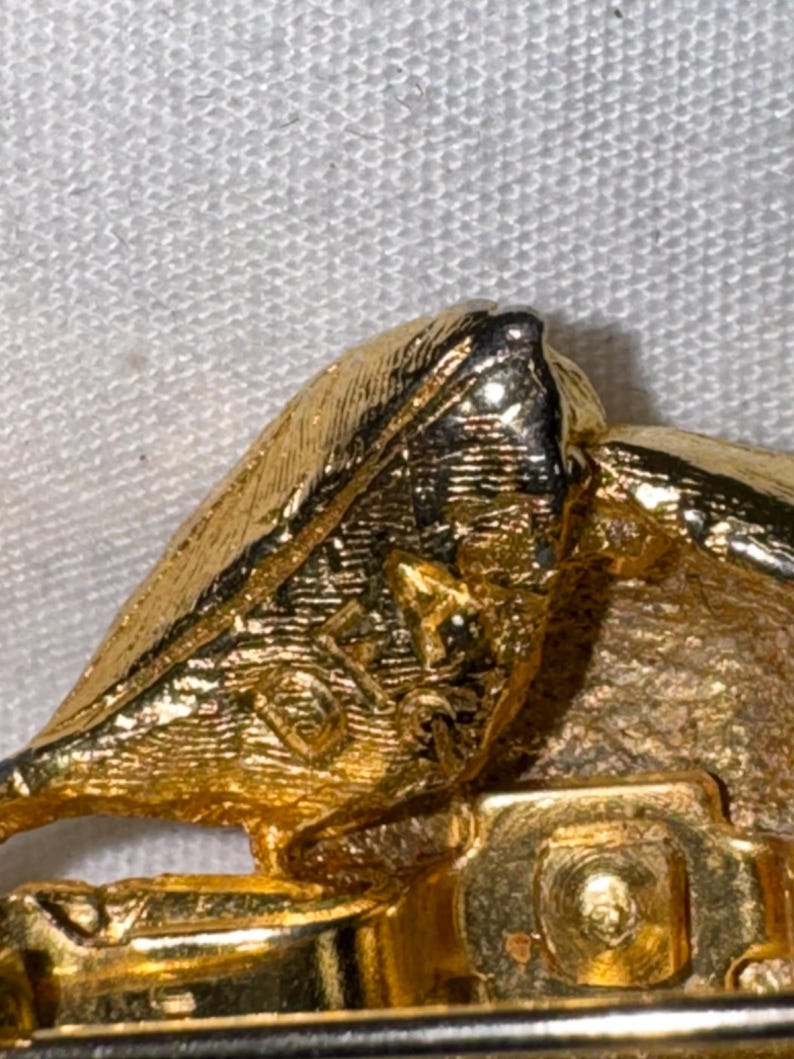 K&ouml;nnte beinhalten: Eine goldfarbene Metallbrosche mit strukturierter Oberfl&auml;che und einem sichtbaren Stempel mit der Aufschrift 'DORLAN' auf der R&uuml;ckseite, inklusive eines sicheren Nadelmechanismus an der Basis.