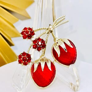 May include: A gold-tone brooch featuring two red strawberry-shaped charms with textured surfaces and gold leaves, accented by three small flower-shaped clusters of bright red rhinestones on twisted gold stems.