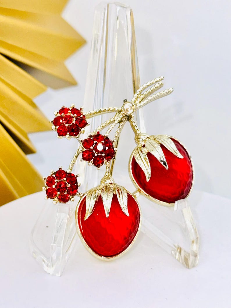 May include: A gold-tone brooch featuring two red strawberry-shaped charms with textured surfaces and gold leaves, accented by three small flower-shaped clusters of bright red rhinestones on twisted gold stems.