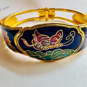 Può includere: Un bracciale rigido incernierato color oro con un pannello centrale in smalto blu scuro caratterizzato da un design a farfalla multicolore e fogliame verde. La struttura in metallo presenta un bordo smerlato e una chiusura sicura con fili d'oro.