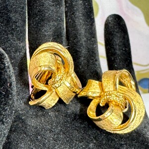May include: A pair of gold-toned clip-on earrings. Each earring features a textured, ribbon-like design with a knotted center. The earrings are displayed on a black surface.