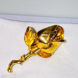 K&ouml;nnte beinhalten: Eine goldfarbene Metallbrosche in Form einer Rosenknospe an einem Stiel mit einem einzelnen Blatt. Die Oberfl&auml;che der Blume und des Stiels hat eine texturierte, reflektierende Oberfl&auml;che, die das Licht auf den geschwungenen Bl&uuml;tenbl&auml;ttern einf&auml;ngt.